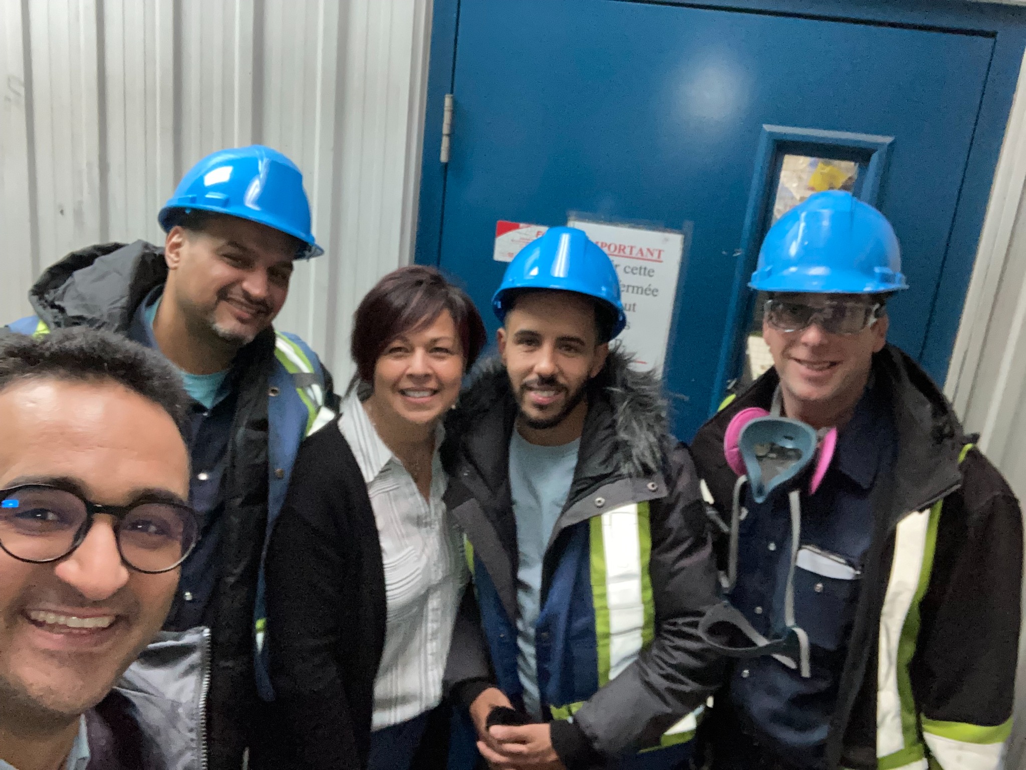 Cinq personnes portant casques et vêtements de sécurité, réunies à l’entrée d’un site industriel.