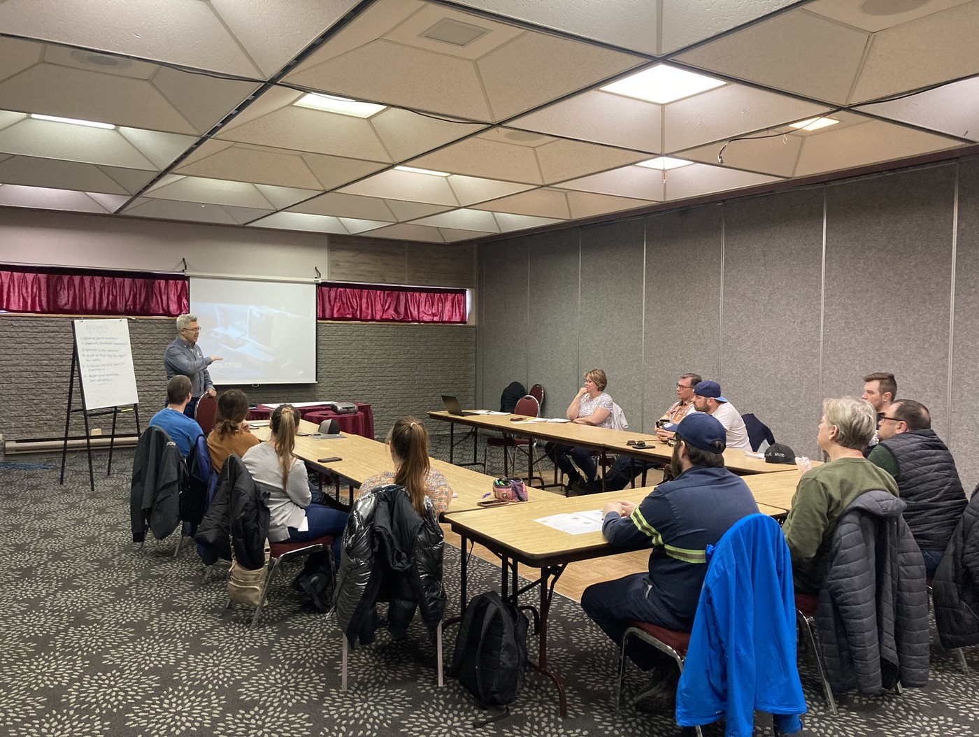 Formateur de Mosaïca Conseils animant une formation devant un groupe de gestionnaires assis en U, dans une salle de conférence avec écran de projection et tableau à feuilles.
